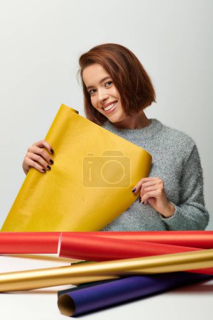 femme souriante en pull confortable tenant papier cadeau jaune sur fond gris, Joyeux Noël concept