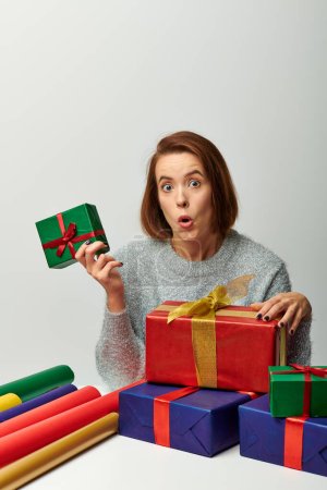 wow emotion, woman in winter sweater holding Christmas present near colorful gift paper on grey