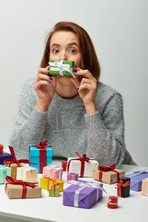 funny woman in sweater holding tiny Christmas present near face and colorful wrapped gifts on grey