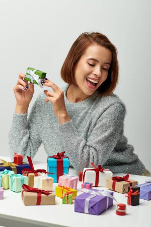 thrilled woman in cozy sweater holding tiny Christmas present near colorful wrapped gifts on grey