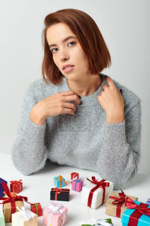 Foto de Hermosa mujer en suéter de invierno sentado entre manojo de regalos de Navidad en la mesa, fondo gris - Imagen libre de derechos
