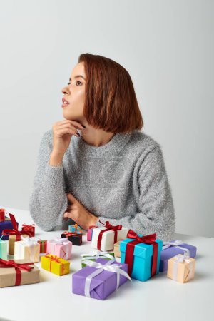 Photo for Dreamy woman in winter sweater fantasizing among bunch of Christmas gifts on table, grey backdrop - Royalty Free Image
