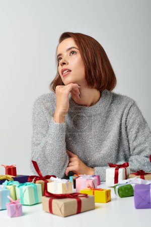 Photo for Pretty woman in winter sweater fantasizing among bunch of Christmas gifts on table, grey backdrop - Royalty Free Image