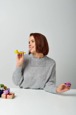 fröhliche Frau im Pullover fantasiert und hält winziges Weihnachtsgeschenk und gelbes Partyhorn auf grau