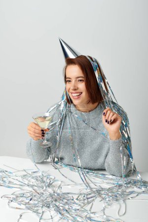 Frau in Partymütze mit silbernem Lametta auf dem Kopf hält Glas Champagner auf grau, Frohes Neues Jahr