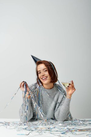 happy woman in party cap with tinsel on head holding glass of martini on grey, Merry Christmas