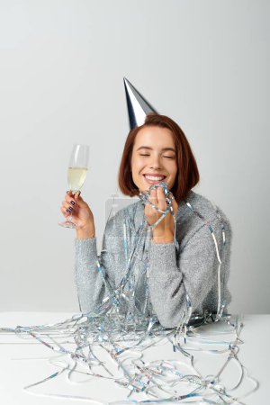 glückliche Frau mit Partymütze und Lametta auf dem Kopf, die ein Champagnerglas hält, während sie einen Neujahrswunsch äußert
