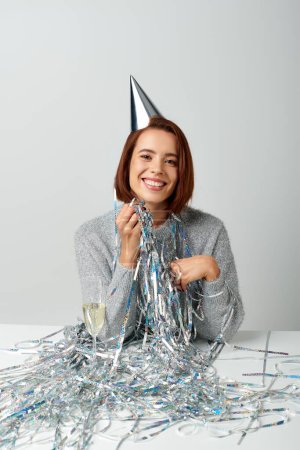 contented woman in party cap and tinsel smiling near champagne glass while celebrating New year