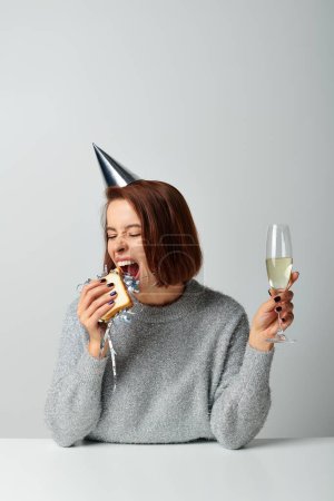 Frau in Partymütze beißt Sandwich mit Lametta und hält Champagnerglas in der Hand und feiert Neujahr