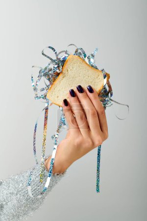abgeschnittene weibliche Hand hält Sandwich mit glänzendem Lametta auf grauem Hintergrund, Happy New Year Konzept