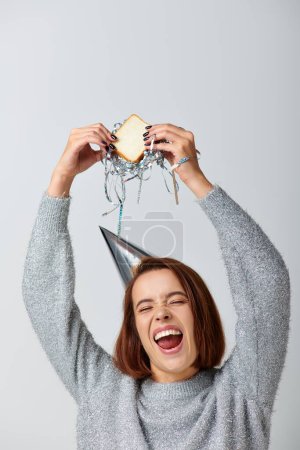 excited woman in party cap holding sandwich with tinsel above head on grey, celebrating New year