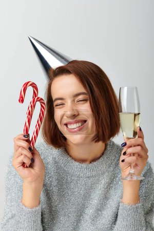Téléchargez les photos : Femme heureuse dans le chapeau de fête tenant verre de champagne et cannes de bonbons sur gris, Joyeux Noël concept - en image libre de droit