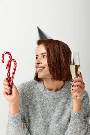 Téléchargez les photos : Femme heureuse dans le chapeau de fête tenant verre de champagne et cannes de bonbons sur gris, Joyeux Noël concept - en image libre de droit
