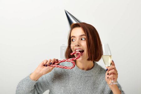Téléchargez les photos : Femme heureuse dans le chapeau de fête tenant verre de champagne et canne à bonbons mordant sur gris, Joyeux Noël - en image libre de droit