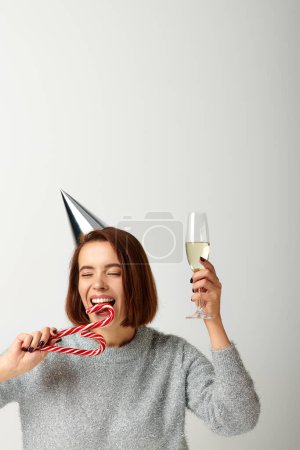 Téléchargez les photos : Jeune femme dans le chapeau de fête tenant verre de champagne et canne à bonbons mordant sur gris, Joyeux Noël - en image libre de droit