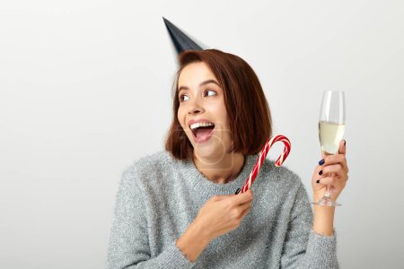 Téléchargez les photos : Femme excitée dans le chapeau de fête tenant verre de champagne et canne à bonbons sur gris, Joyeux Noël - en image libre de droit