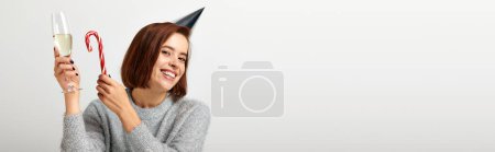 positive woman in party cap holding champagne glass and candy cane on grey, Merry Christmas, banner