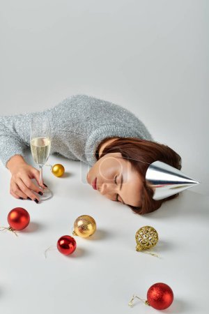 woman in party cap sleeping on table near champagne glass and Christmas baubles on grey backdrop