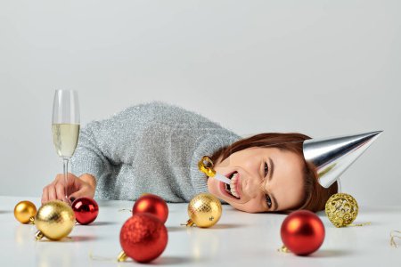 woman in party cap lying on table near champagne glass and Christmas baubles, blowing party horn