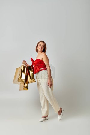 New Year shopping, happy young woman in stylish attire holding shopping bags on grey background