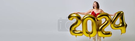 Banner de Año Nuevo, mujer joven emocionada con atuendo de moda sosteniendo globos con números 2024 en gris