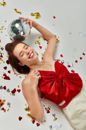 New Year party, young cheerful woman with disco ball lying on floor near confetti on grey backdrop