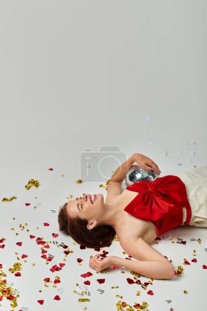 Fiesta de Año Nuevo, mujer joven emocionada con bola disco tirada en el suelo cerca de confeti sobre fondo gris