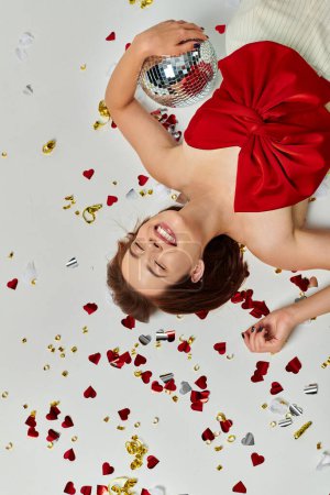 Año Nuevo, mujer emocionada con bola de discoteca tirada en el suelo cerca de confeti sobre fondo gris, vista superior
