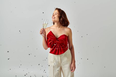 Happy New Year, cheerful woman in party attire holding glass of champagne near confetti on grey