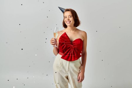 Happy New Year, cheerful woman in party cap holding glass of champagne near confetti on grey