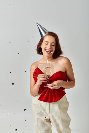 Frohes neues Jahr, positive junge Frau mit Partymütze hält ein Glas Champagner in der Nähe von Konfetti auf grau