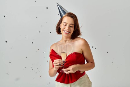 Feliz Navidad, mujer joven positiva en la tapa del partido sosteniendo la copa de champán cerca de confeti en gris