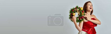 Téléchargez les photos : Bannière de la saison des fêtes, femme excitée avec un maquillage chatoyant tenant une couronne de Noël sur fond gris - en image libre de droit