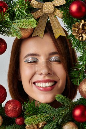 Téléchargez les photos : Vue rapprochée de la femme heureuse avec un maquillage chatoyant souriant à l'intérieur du cadre de couronne de Noël décorée - en image libre de droit