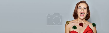 Merry Christmas banner, excited woman in red attire with bows all over her body on grey backdrop