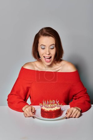 aufgeregte Frau in roter Kleidung blickt auf Bento-Torte mit Happy Birthday Kerzen auf grauem Hintergrund