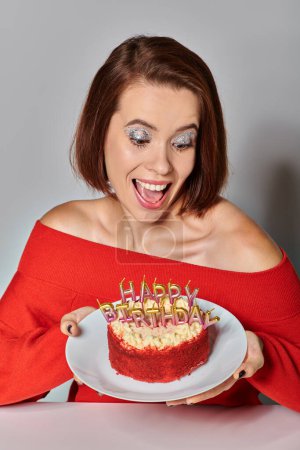 thrilled woman in red attire looking at bento cake with Happy Birthday candles on grey background