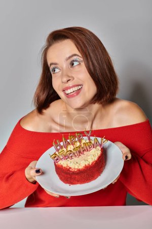 erstaunt Frau in roter Kleidung hält Bento-Kuchen mit Happy Birthday Kerzen auf grauem Hintergrund