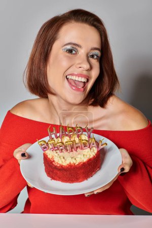 mujer feliz recortado en traje rojo cerca de pastel de bento con velas de feliz cumpleaños sobre fondo gris