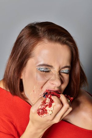 hungry birthday girl with closed eyes biting delicious piece of red velvet cake on grey background