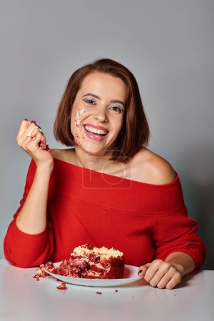chica de cumpleaños positiva con la cara sucia comiendo delicioso pastel de terciopelo rojo sobre fondo gris