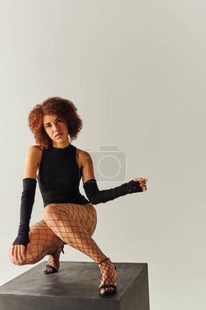 attraktive junge afrikanisch-amerikanische Frau in sexy schwarzer Kleidung hockt auf schwarzem Würfel, Modekonzept