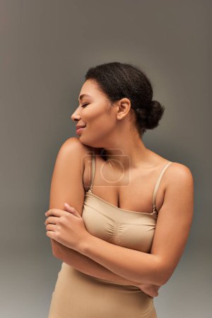 joyous beautiful african american woman in pastel underwear posing on gray backdrop, fashion concept