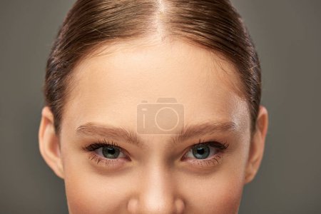 Ausgeschnittene Ansicht einer jungen Frau mit blauen Augen und perfekter Haut, die in die Kamera auf grauem Hintergrund blickt