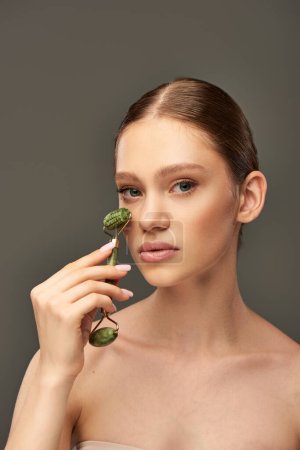 young woman with perfect skin massaging face with jade roller on grey background, skin rejuvenation