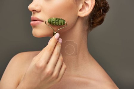 mujer recortada con perfecta piel masajeando la cara con rodillo de jade sobre fondo gris, cuidado de la piel