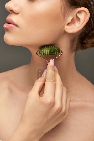 cropped woman with perfect skin massaging face with green jade roller on grey background, skin care