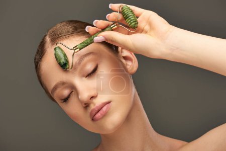 young woman with perfect skin massaging face with jade roller on grey background, pampering skin