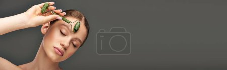 young woman with perfect skin massaging face with jade roller on grey background, banner