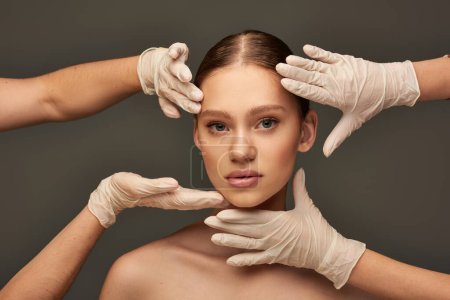 estheticians in medical gloves examining face of young woman on grey background, facial treatment